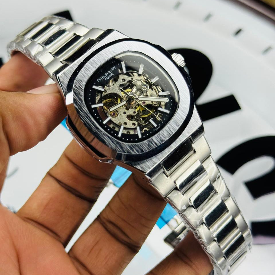 Silver wristwatch with a visible mechanism held by a hand, blurred clock face in the background